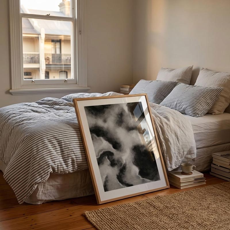 A framed black and white abstract art print with a light wood frame leans against a bed in a softly lit bedroom. The room features neutral beige walls, striped bedding, a window with daylight, and a woven rug on the wooden floor.