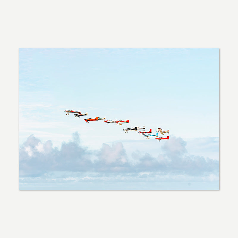A rectangular art print showing a formation of vintage aerobatic airplanes flying across a pale blue sky with soft clouds below, presented on a light gray background.