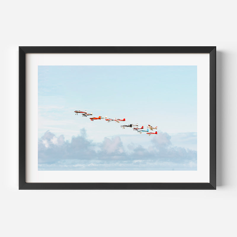 Framed wall art featuring a formation of vintage airplanes flying across a pale blue sky with soft clouds below, shown in a black frame against a light gray background.