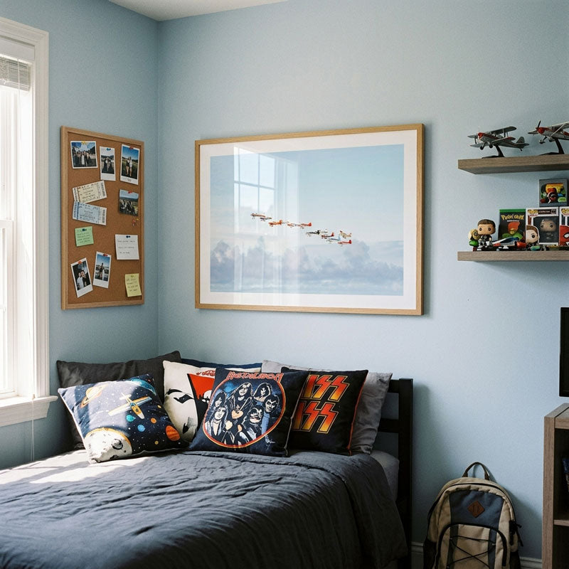 A styled bedroom corner with a bed dressed in dark bedding and several decorative pillows, beneath a large framed print of fighter jets flying over clouds. The room has light blue walls, a cork bulletin board, wooden shelves with toy models and figurines, and a backpack on the floor.