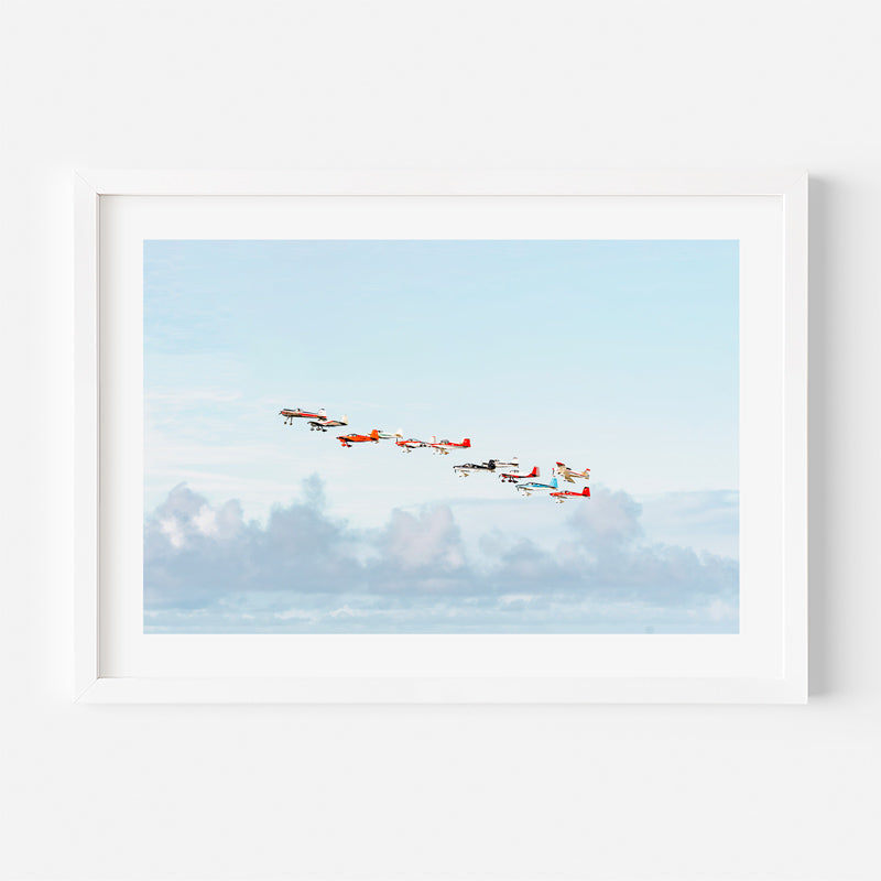 A framed art print featuring a formation of small red and white airplanes flying across a light blue sky with soft clouds, displayed on a clean white wall.