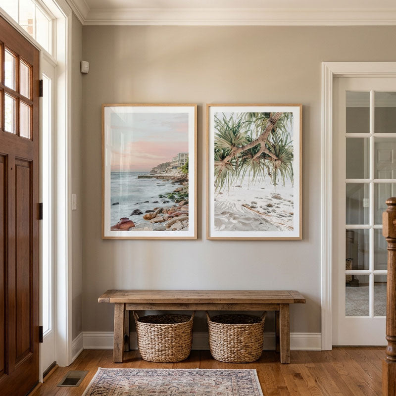 Two framed landscape art prints hang on a beige wall above a rustic wooden bench with two woven baskets in a home entryway. The left print shows a rocky coastal shoreline at sunset, and the right print shows a beach scene with pine branches.
