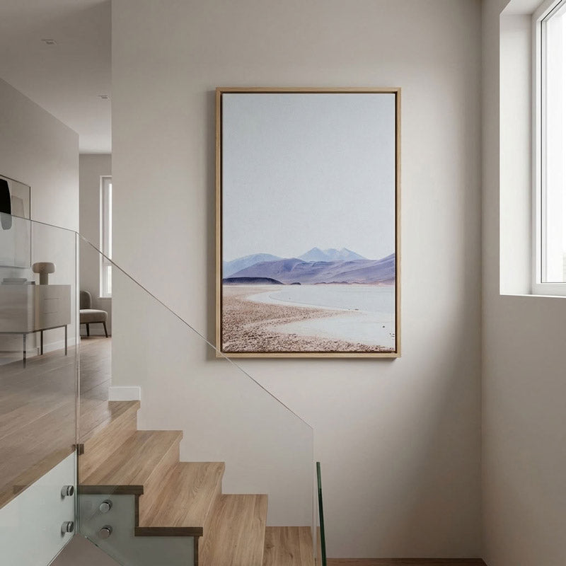 A vertical framed art print depicting a minimalist beach and mountain landscape, styled on a neutral beige wall above a wooden staircase in a modern home interior.