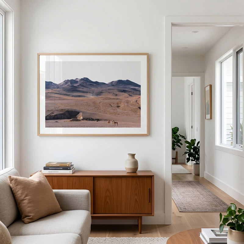 A framed landscape art print of a desert scene with mountains hangs on a white wall in a modern living room setting above a wooden sideboard.