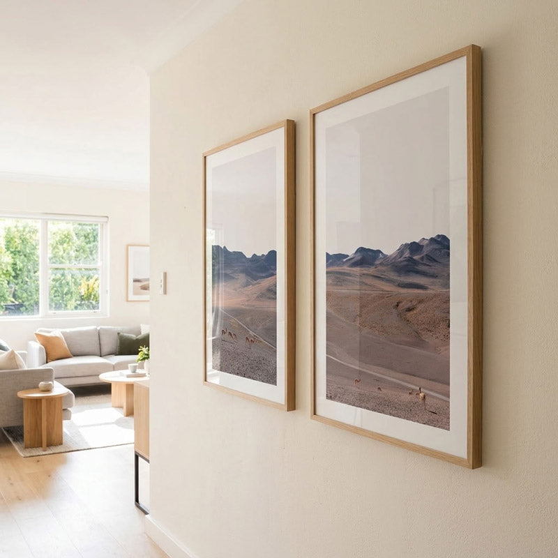 Two vertical framed landscape art prints with light wood frames and white mats hanging on a beige wall in a modern living room setting.