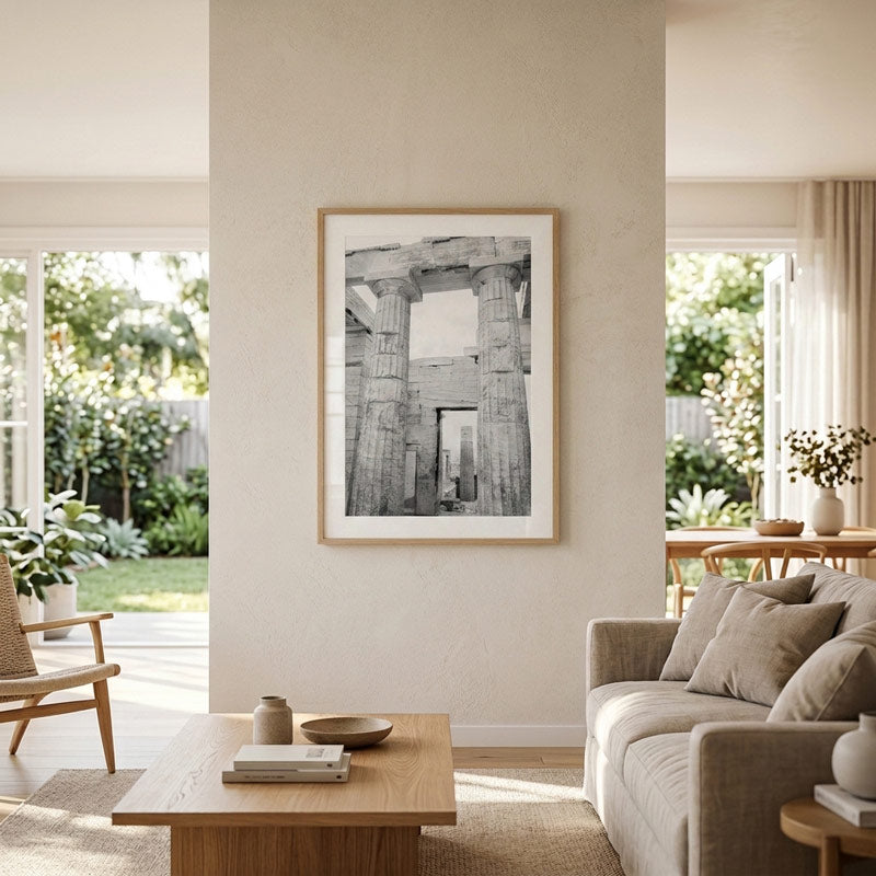 A framed black and white architectural art print of ancient stone columns hangs on a beige wall in a modern, sunlit living room with a sofa, wooden coffee table, and large windows overlooking a garden.