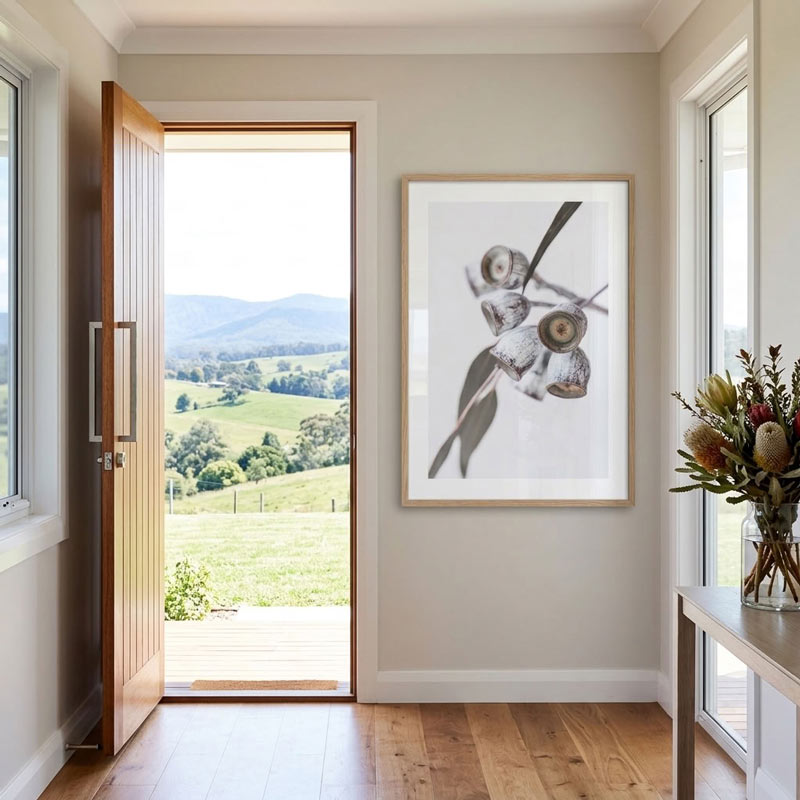 A framed botanical art print of a eucalyptus branch with gum berries hangs on a light beige wall in a modern entryway with a wooden door open to a green valley landscape.