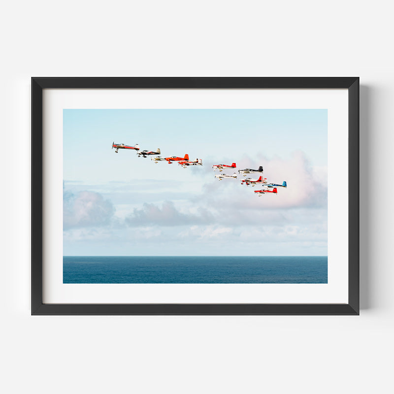 Framed wall art featuring a photograph of six vintage aircraft flying in formation over a blue ocean under a pale sky, shown in a black frame with a white mat against a light gray background.