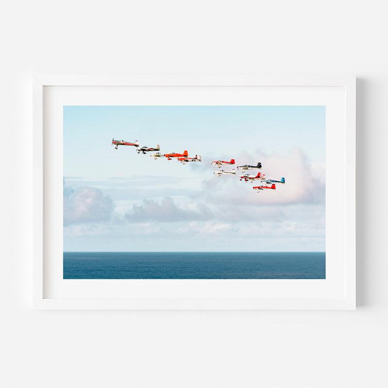 A framed art print featuring a formation of vintage red and white airplanes flying over a blue ocean under a light sky, displayed on a clean white wall.