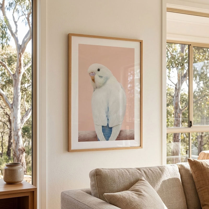 A framed art print featuring a white cockatoo against a soft pink background, hanging on a beige wall above a sofa in a bright, modern living room.
