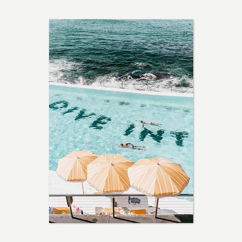 People swimming in a pool with ocean view and 'Dive In' text, surrounded by beach umbrellas.