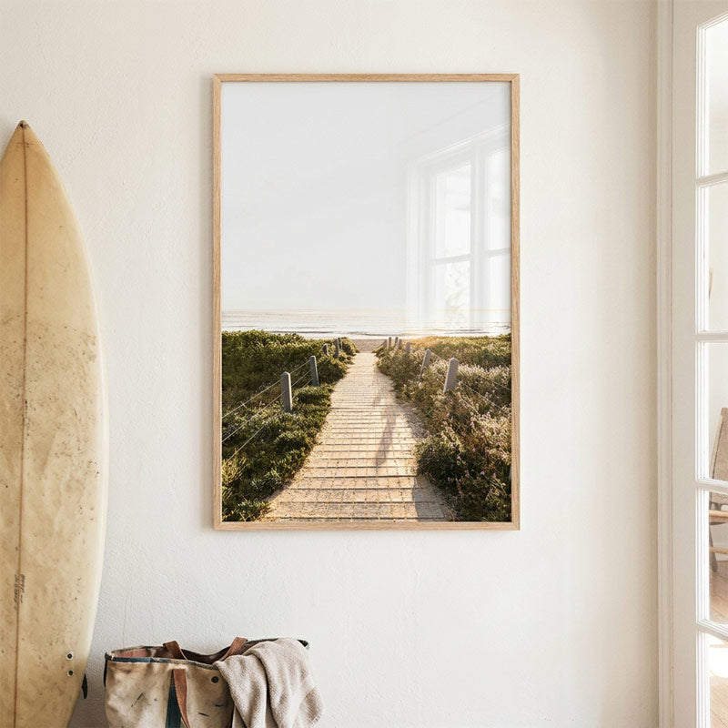 Framed artwork of a beach scene with a wooden path on a white wall.