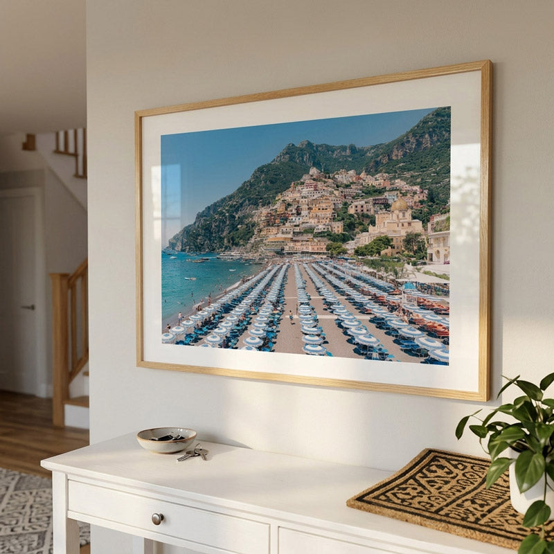 A framed photographic print of a coastal town with beach umbrellas and blue water, displayed on a white wall above a console table in a bright interior room.
