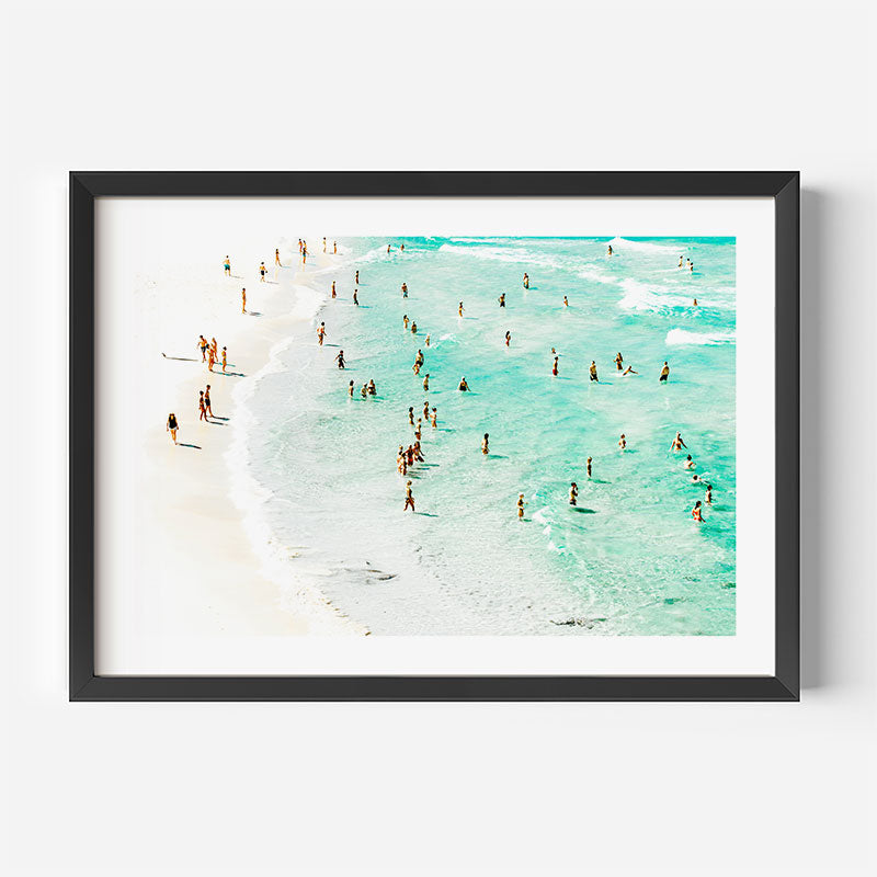 Framed artwork of a beach scene with people in the water on a white wall.