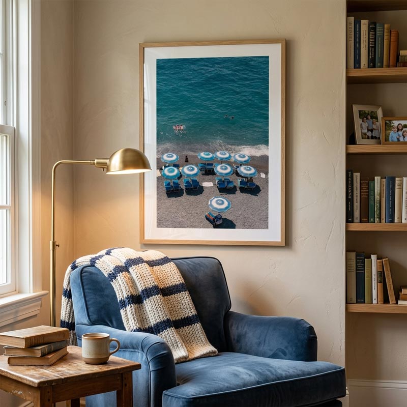 A framed beach photograph hanging on a beige wall above a blue armchair in a styled living room. The print shows blue lounge chairs on a sandy beach with turquoise ocean water.