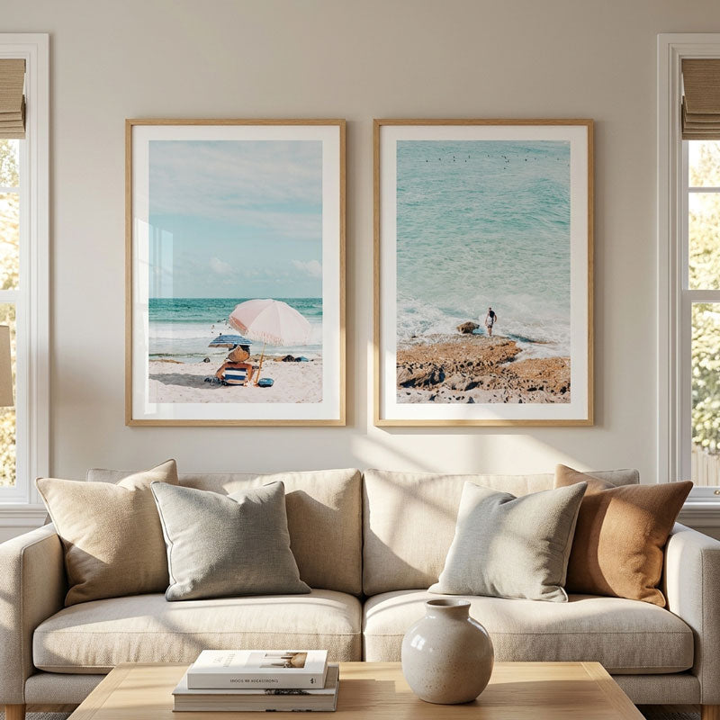 Two framed beach art prints hanging side-by-side on a beige wall above a light beige sofa with neutral-toned pillows in a bright, modern living room.