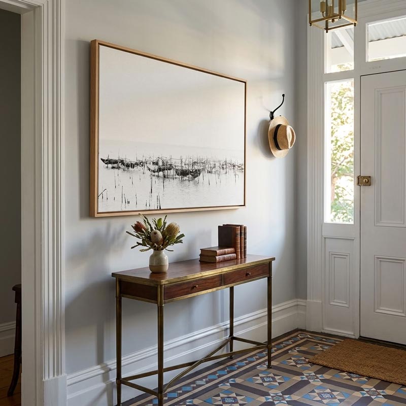 A large framed black and white abstract landscape print hangs on a light gray wall in a bright entryway above a wooden console table with brass legs. A hat hangs on a hook, books sit on the table, and a patterned tile floor is visible.