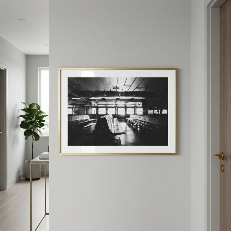 A framed black and white photograph of an empty interior space with benches and windows, displayed on a light gray wall in a modern hallway with a plant and doorway visible.