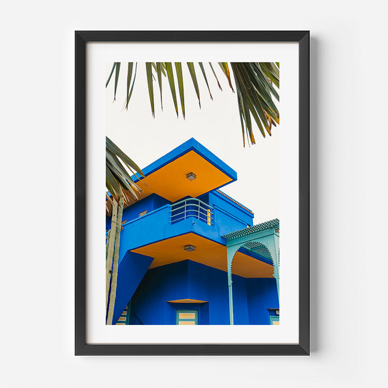 Framed photograph of a colorful building with palm leaves on a white wall