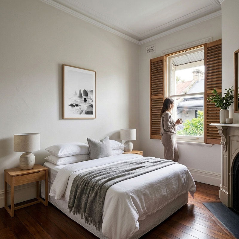 A woman stands by a window in a bright, modern bedroom with a neatly made bed, wooden nightstands, and warm brown shutters. The room has light walls, hardwood floors, and a fireplace on the right.