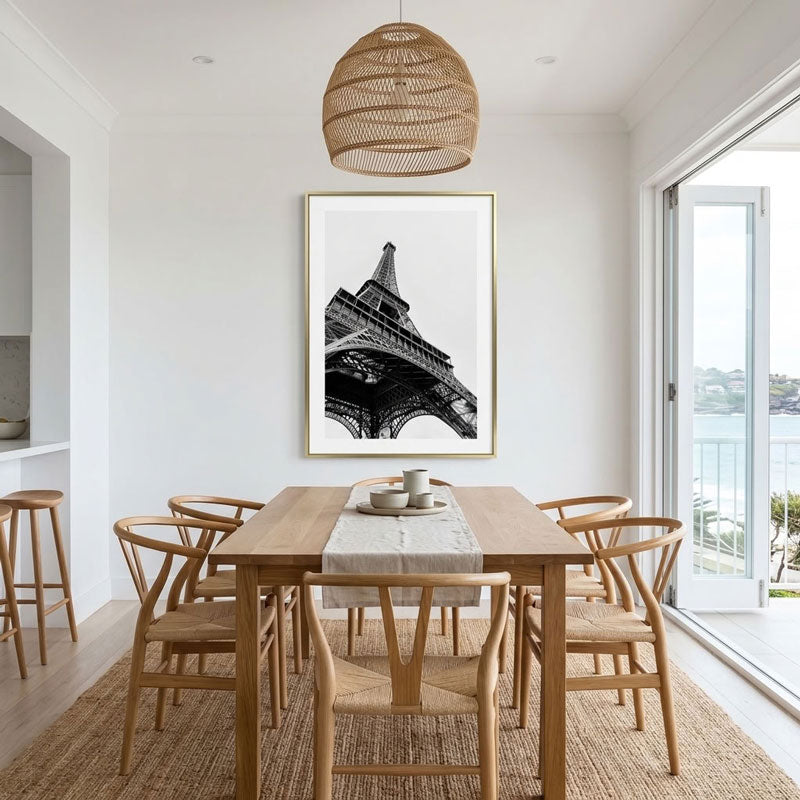 A framed black and white Eiffel Tower art print hangs on a white wall above a wooden dining table and chairs in a bright, modern dining room with a woven pendant light and large windows.