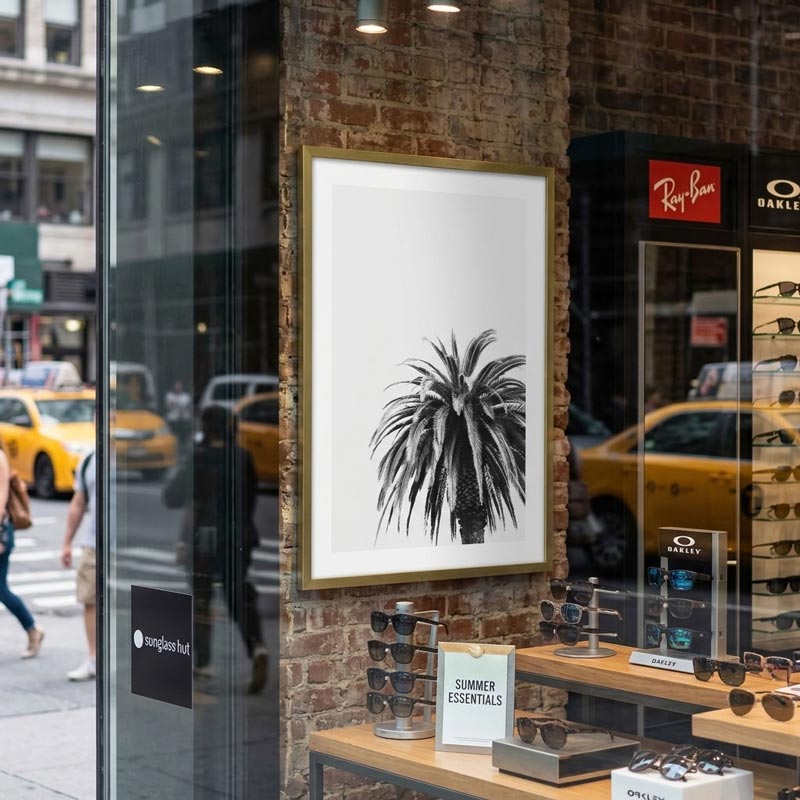 A framed black and white palm tree art print with a gold frame hanging on a brick wall in a retail store window display. Yellow taxis and pedestrians are visible in the street reflection, with eyeglasses displayed on stands below.