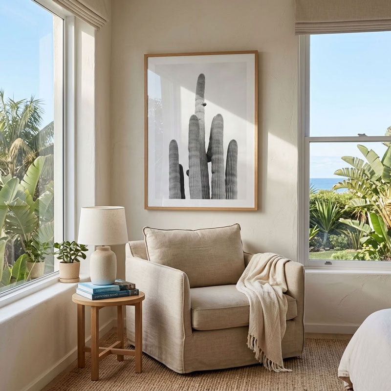 A framed black and white cactus art print hangs on a beige wall above a matching beige armchair in a bright, sunlit room with windows, plants, and a side table.