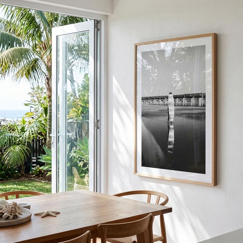 A framed black and white landscape photograph hanging on a white wall in a bright dining room with a wooden table and chairs, with an open glass door showing tropical greenery outside.