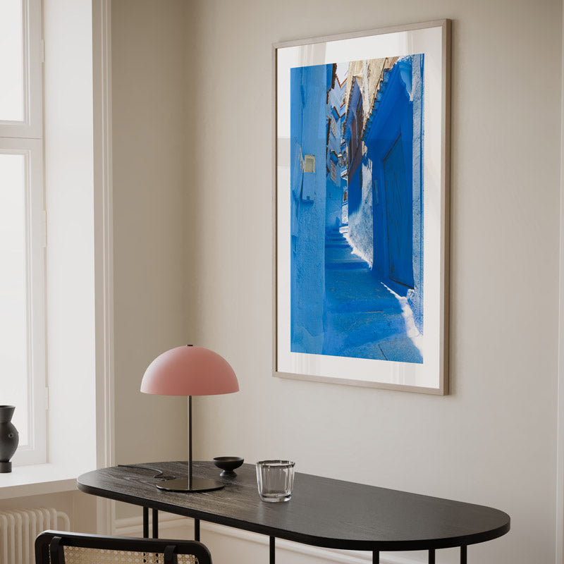 Framed artwork of a blue alleyway on a wall above a black table with a pink lamp.