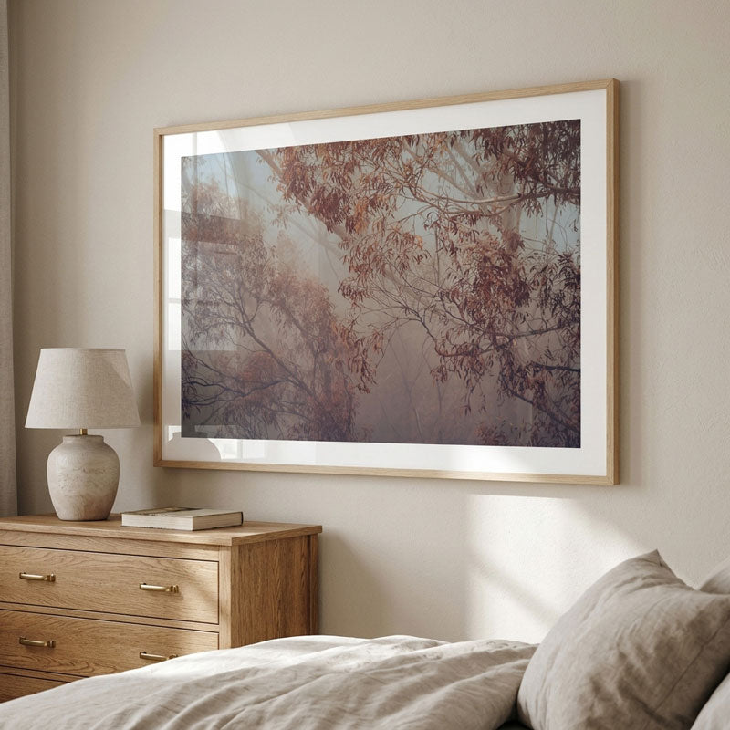 A large framed art print depicting a misty forest with autumn leaves, styled in a bedroom setting above a wooden nightstand and bed.