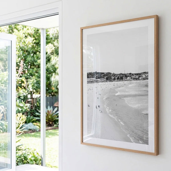 A framed black and white beach photograph hanging on a white wall next to a glass door that looks out onto a green garden.