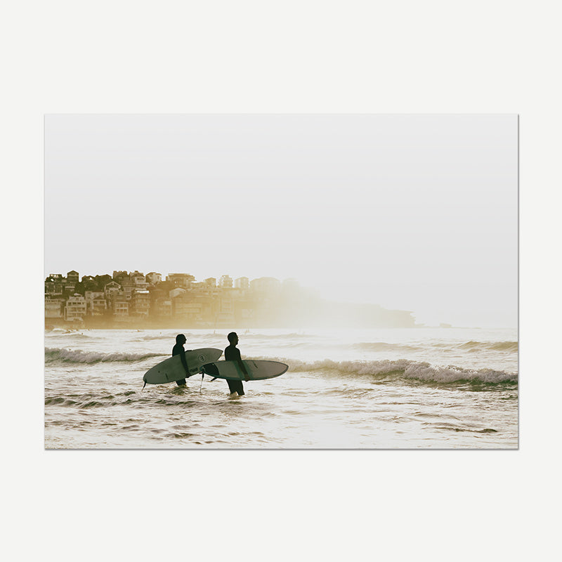 A square photographic art print depicting two surfers walking into the ocean with their boards, silhouetted against a hazy, bright sky with a coastline and buildings in the background.