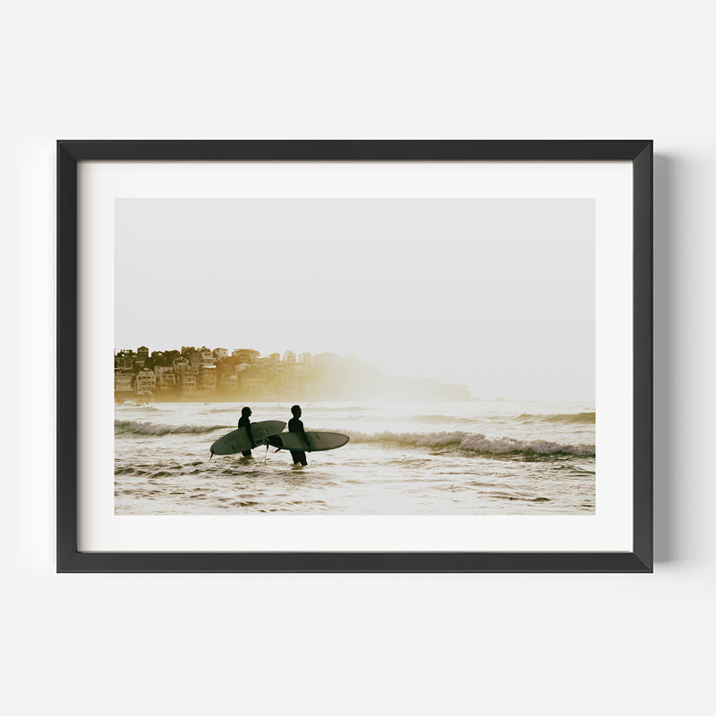 A framed photographic art print of two surfers carrying boards at the beach during a misty, golden hour, presented in a black frame with a white mat against a light gray background.