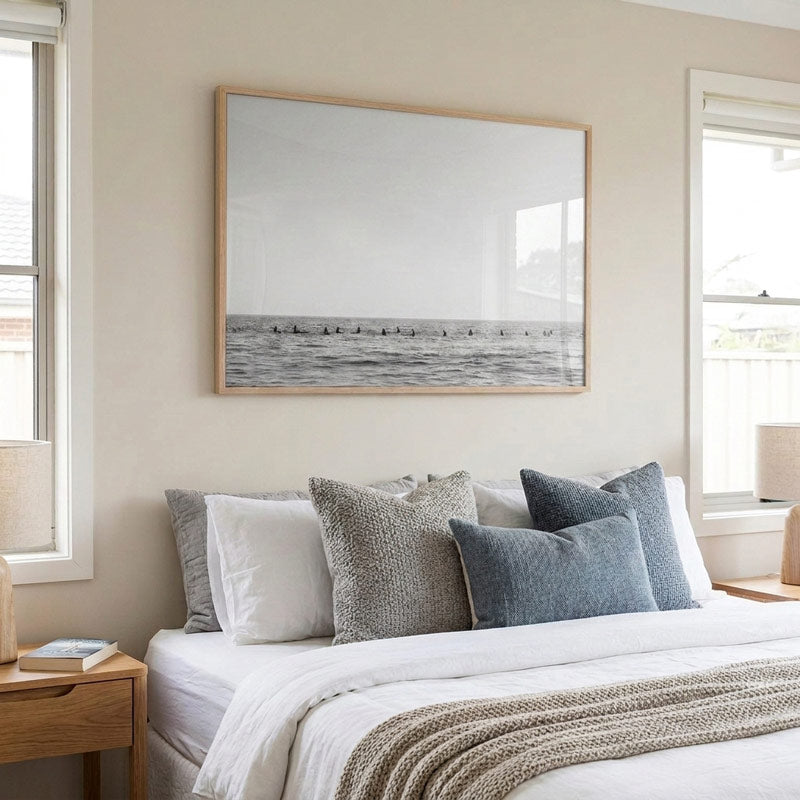 A large framed black and white ocean photograph hangs on a beige wall above a neatly made bed with white linens, textured gray pillows, and a beige knit throw blanket.