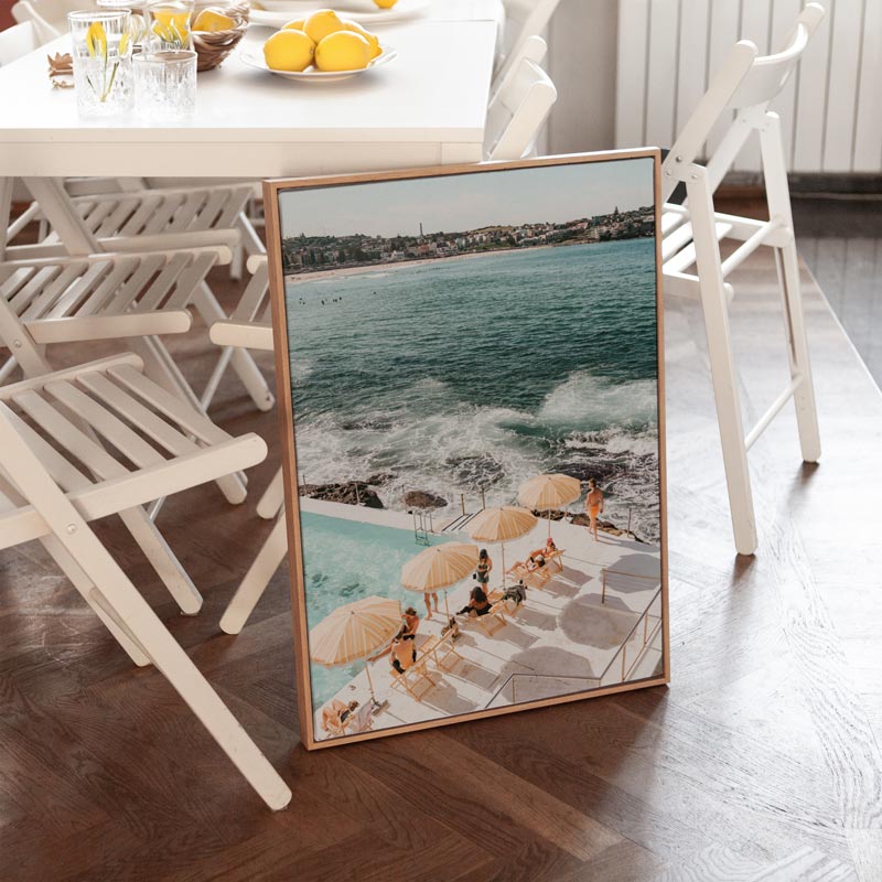 Framed artwork of a beach scene on a wooden floor with a dining table and chairs in the background.