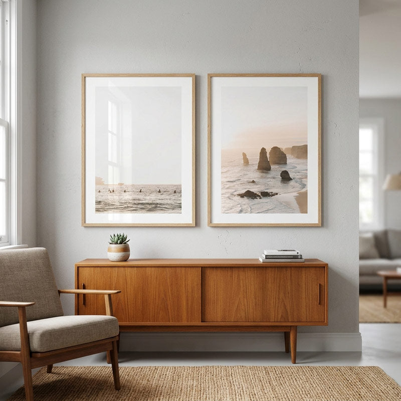 Two framed minimalist beach and rock formations art prints hanging on a light gray wall above a mid-century modern wooden sideboard in a styled living room.