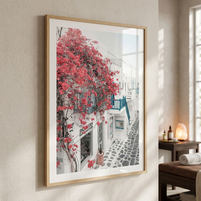 A framed art print depicting a white Mediterranean-style street with vibrant pink bougainvillea flowers, hanging on a textured beige wall in a spa-like room.