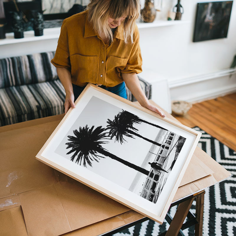 Person holding a framed black and white artwork of palm trees in a living room.