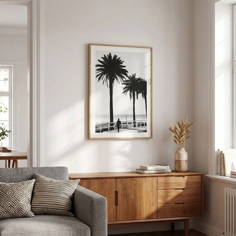 A framed black and white art print of palm trees on a beach, displayed on a light gray wall above a mid-century modern wooden sideboard in a bright, modern living room.
