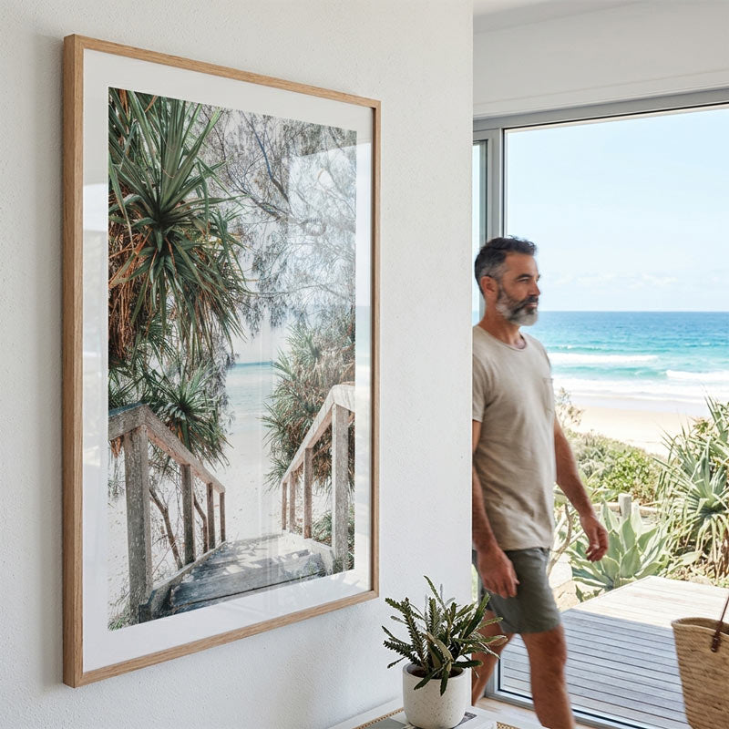 Man standing by a large window with a scenic view of the ocean, next to a framed beach photo on the wall.