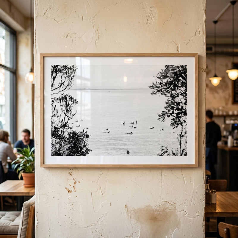 A framed black and white landscape photograph of a lake with trees and small birds, displayed on a textured cream wall in a cafe setting.