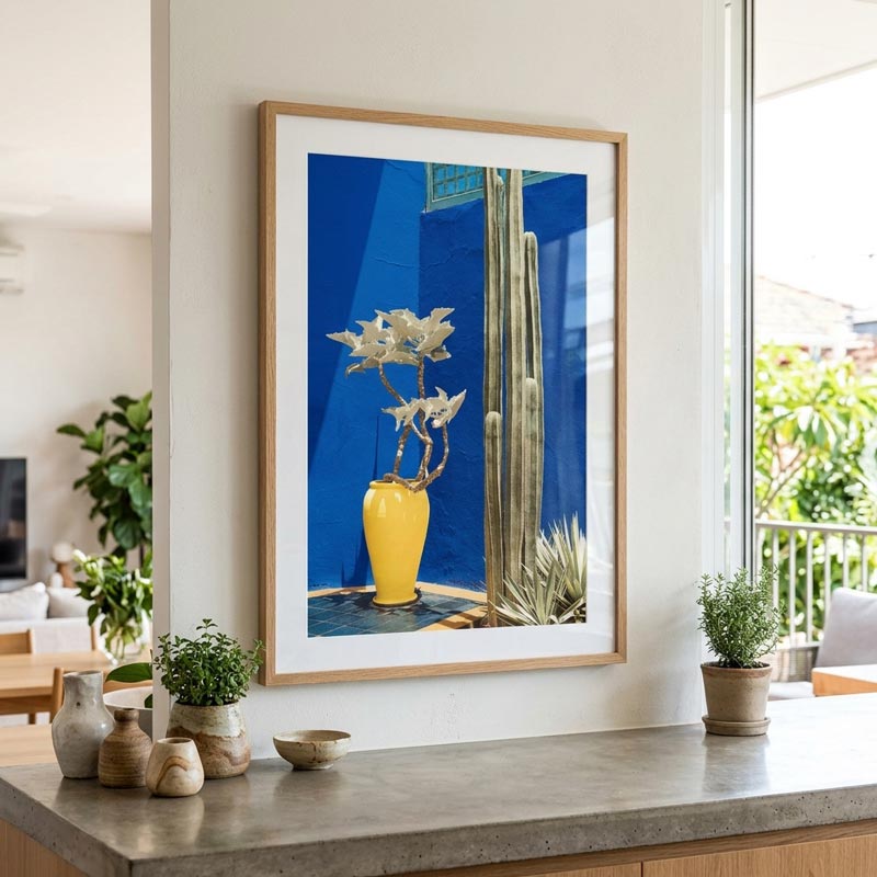 A framed art print featuring a bright yellow vase with white flowers against a vivid blue background, displayed on a light-colored wall above a stone countertop with small plants and decorative vases.