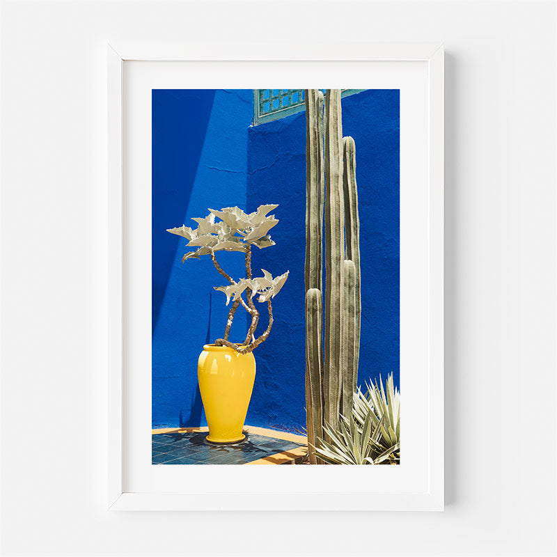 Framed photograph of a yellow vase with a plant against a blue wall.