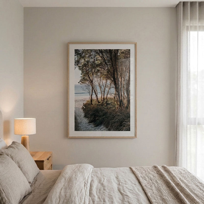 A framed landscape art print depicting a forest path with trees and a beach in the distance, styled on a neutral beige bedroom wall above a bed with neutral-toned bedding.