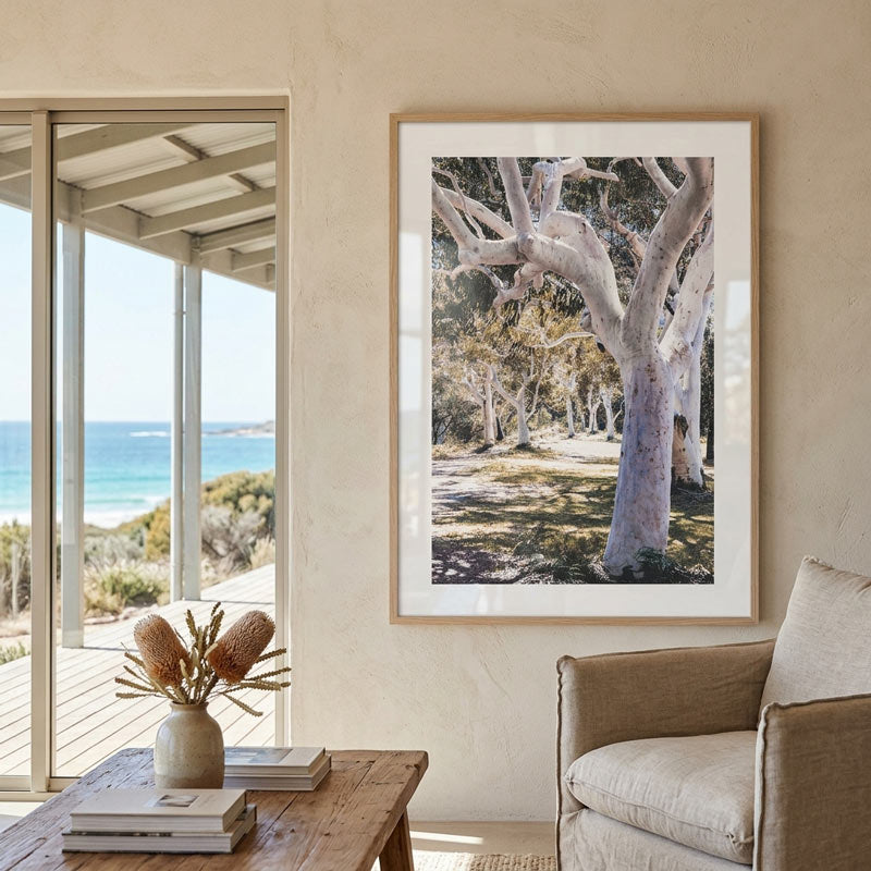 A framed art print depicting a coastal tree-lined path with a beach and ocean in the background, styled in a modern living room setting next to a window and an armchair.