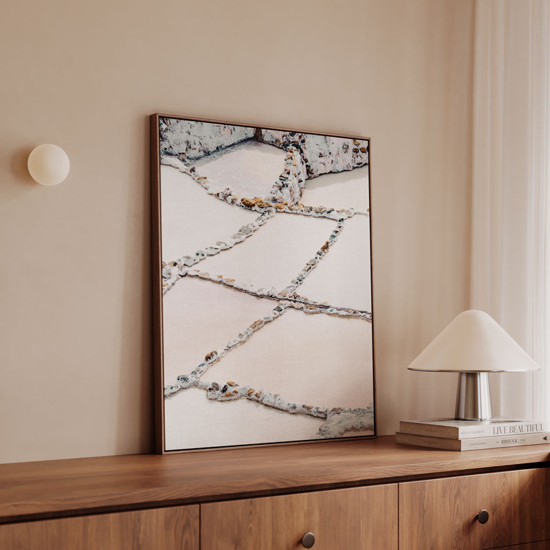 Framed abstract artwork on a wall above a wooden cabinet with a lamp and books.