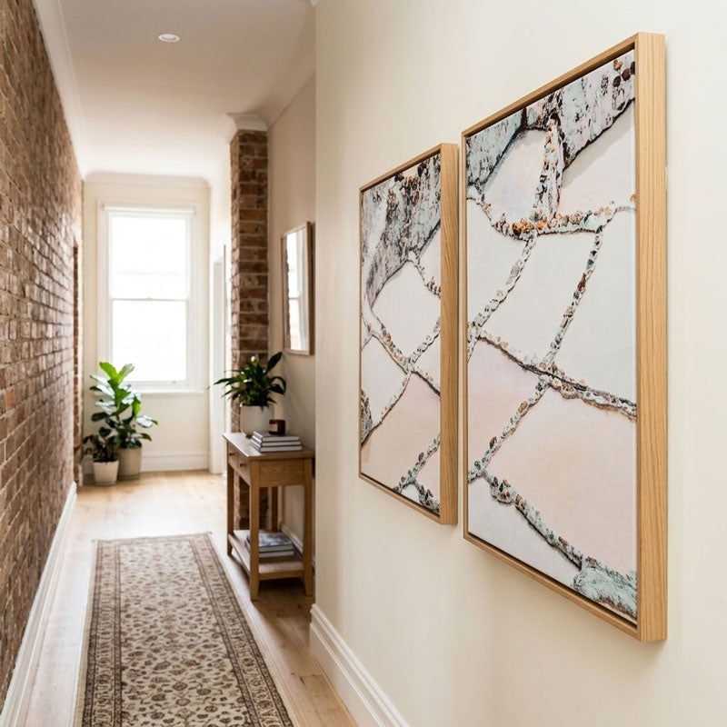 Two framed abstract wall art prints hang on a cream hallway wall next to a brick column and wooden console table with plants.