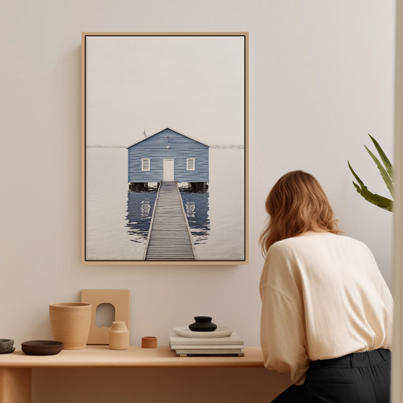 Person looking at a framed artwork of a blue house and dock on a wall.