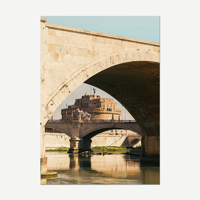 A vertical photographic print of a stone arch bridge over water, with a historic fortress visible in the background under a pale blue sky.