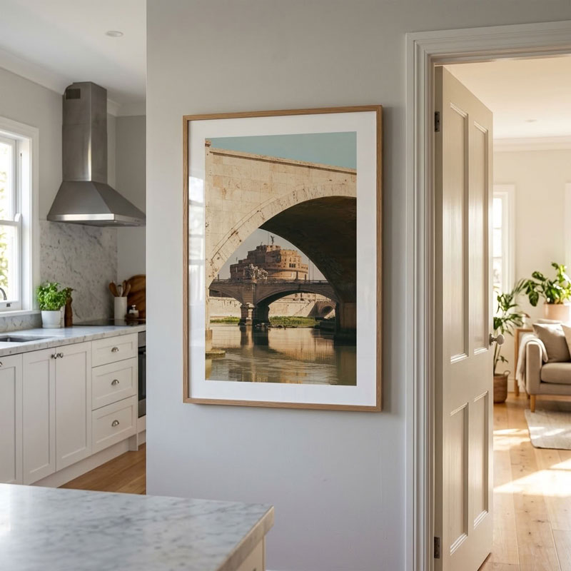 A framed art print depicting an old stone bridge over water with a castle, displayed on a light gray wall in a modern kitchen and living room interior.